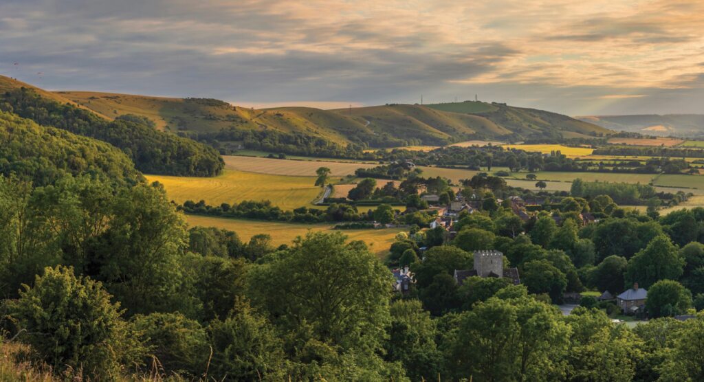 Panoramic view of the West Sussex countryside at sunset, showing rolling green hills, patchwork fields, and a small village nestled among trees in the valley.